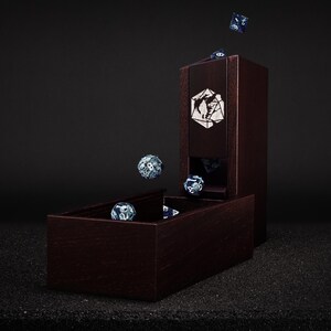 May include: A dark wood dice tower and tray set with a geometric design. Several blue and white dice are shown in the tray, falling from the tower, and in mid-air. The tower has a decorative emblem on the front.