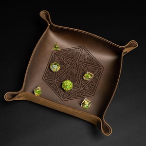 May include: A brown leather dice tray with a geometric design and several multi-sided dice. The tray has a raised edge and stitched details. The dice are clear with green inclusions. The tray is designed for tabletop gaming.