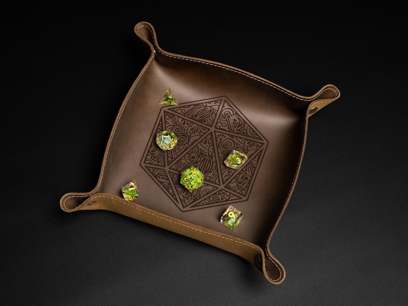 May include: A brown leather dice tray with a geometric design and several multi-sided dice. The tray has a raised edge and stitched details. The dice are clear with green inclusions. The tray is designed for tabletop gaming.