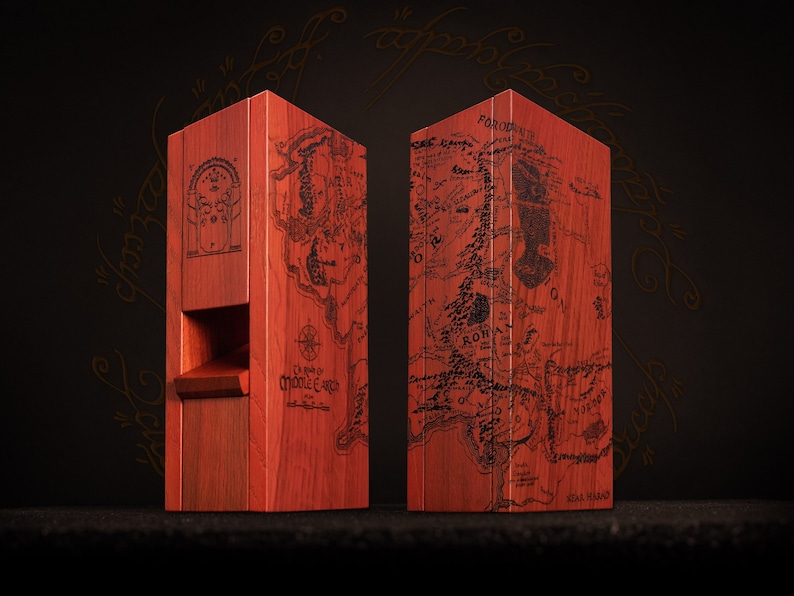 May include: Two reddish-brown wooden boxes, each engraved with a detailed map of Middle-earth in black. One box has a carved architectural design. The boxes are displayed against a dark background, showcasing the intricate map details.