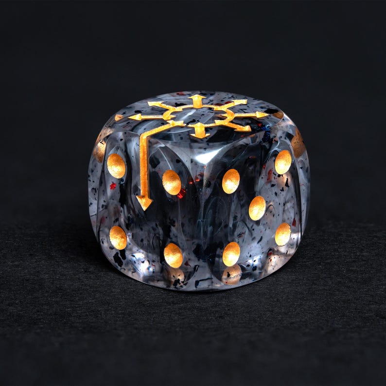 May include: A six-sided, clear resin die with a swirling black and white pattern. The die features gold-colored pips and a gold symbol on the top face. The symbol is a stylized arrow design.