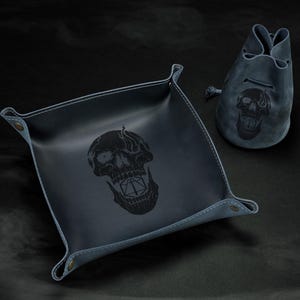 May include: A dark blue leather dice tray and drawstring bag set. Both items feature a black skull design. The square tray has snap closures at the corners. The bag has a drawstring closure. The set is designed for tabletop gaming.