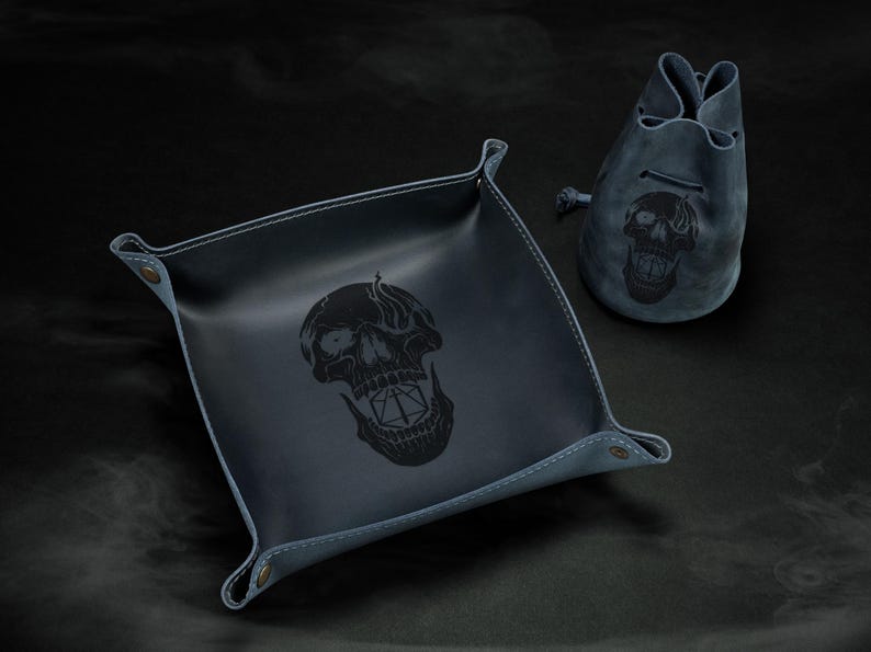 May include: A dark blue leather dice tray and drawstring bag set. Both items feature a black skull design. The square tray has snap closures at the corners. The bag has a drawstring closure. The set is designed for tabletop gaming.