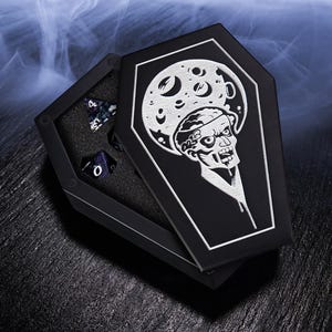 K&ouml;nnte beinhalten: Schwarze Sarg-W&uuml;rfelbox mit einem Zombie- und Mond-Design. Die offene Box zeigt mehrere vielseitige W&uuml;rfel. Die Box ist schwarz mit einer wei&szlig;en Grafik. Die W&uuml;rfel sind dunkelblau mit wei&szlig;en Zahlen. Das Thema ist gotisch und Fantasy.