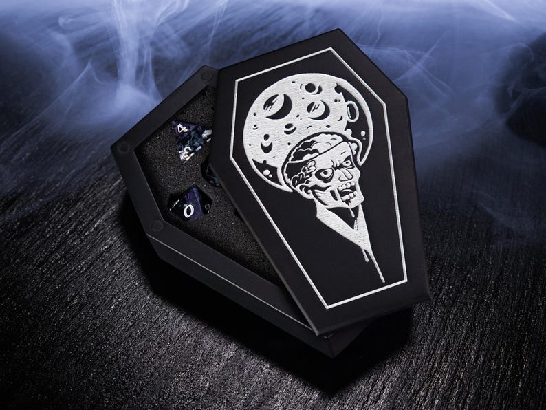 K&ouml;nnte beinhalten: Schwarze Sarg-W&uuml;rfelbox mit einem Zombie- und Mond-Design. Die offene Box zeigt mehrere vielseitige W&uuml;rfel. Die Box ist schwarz mit einer wei&szlig;en Grafik. Die W&uuml;rfel sind dunkelblau mit wei&szlig;en Zahlen. Das Thema ist gotisch und Fantasy.