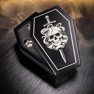 May include: Black coffin-shaped box with a white skull and snake design. The box is open, revealing a small, sparkling object inside. The coffin lid features a sword through the skull. The box is on a dark surface.