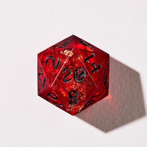 May include: A red, twenty-sided die (D20) with black numerals and gold inclusions. The number 20 is visible on one face. The die is made of transparent resin. Suitable for role-playing games and tabletop gaming.