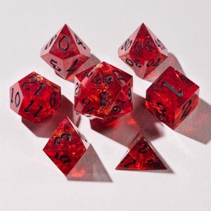 May include: A collection of seven red polyhedral dice with gold flecks and black numbering. The set includes a d20, d12, d10, d8, d6, and d4. These dice are designed for tabletop role-playing games and other dice-based games.