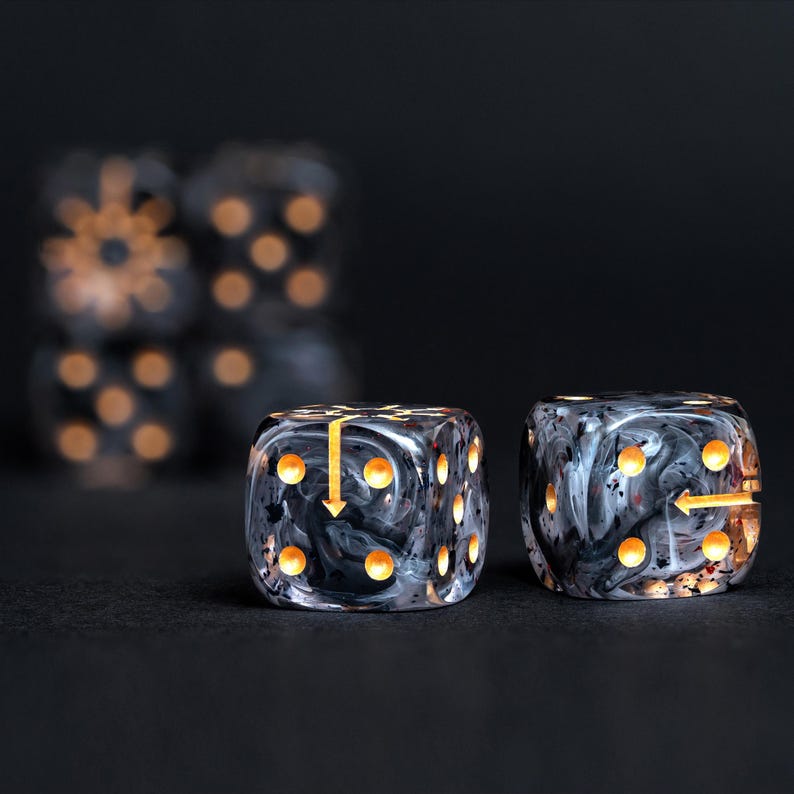 May include: Two dice with a marbled black and white design and gold-colored pips. One die has a gold arrow pointing down, and the other has a gold arrow pointing to the right. The dice are on a black surface.