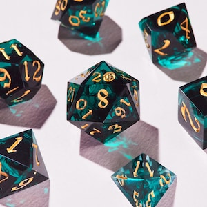 May include: A selection of polyhedral dice with a marbled teal and black design. The dice have gold-coloured numbers and are arranged on a white surface. The dice are for tabletop games.