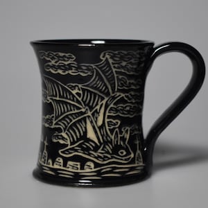 May include: Black ceramic mug with a bat design. The bat is depicted in a cream color, flying against a background of clouds and a landscape. The mug has a curved handle and a slightly flared rim.