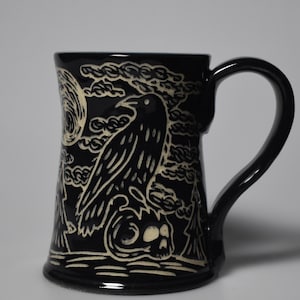 May include: A black ceramic mug featuring a carved, cream-colored graphic design. The illustration depicts a raven perched atop a skull, set against a backdrop of a full moon, swirling clouds, and pine trees. The mug has a curved handle and base.