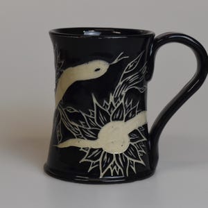 May include: A glossy black ceramic mug featuring a hand-carved sgraffito design. The artwork displays a cream-coloured snake winding through a stylised sunflower motif. The mug has a curved handle and a flared rim, showcasing a high-contrast aesthetic.