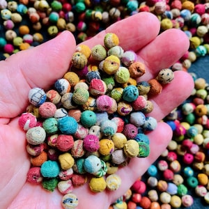 May include: A handful of colorful, fabric-covered beads in various sizes and hues, including red, blue, yellow, and green. The beads are tightly packed together, with a few spilling out of the hand. The background is filled with more beads.