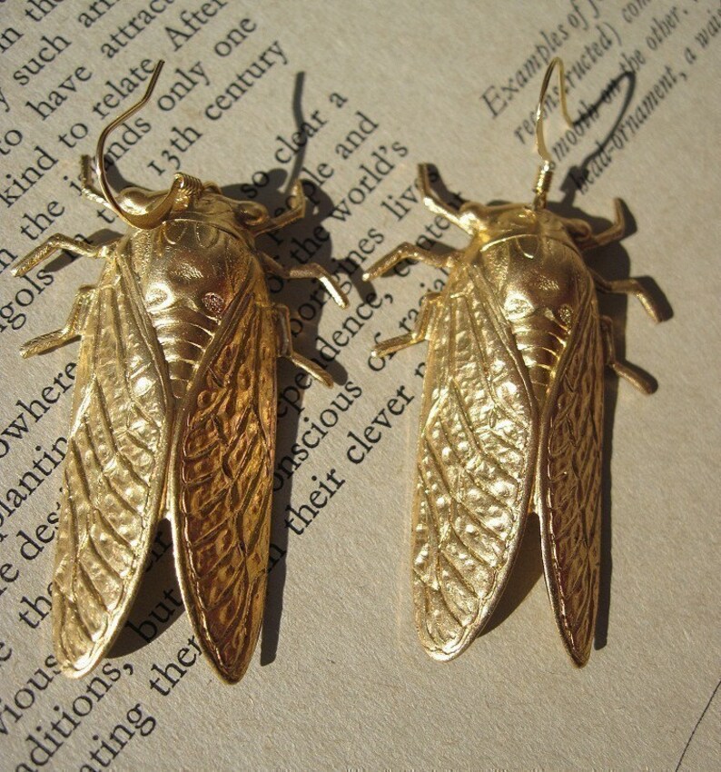 May include: A pair of gold-toned cicada earrings with intricate details. The earrings are made of metal and have a textured finish.