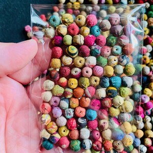 May include: A clear plastic bag filled with a variety of small, colorful fabric-covered beads. The beads are in shades of pink, yellow, green, blue, and red. The background is filled with more of the same beads, creating a vibrant and textured display.