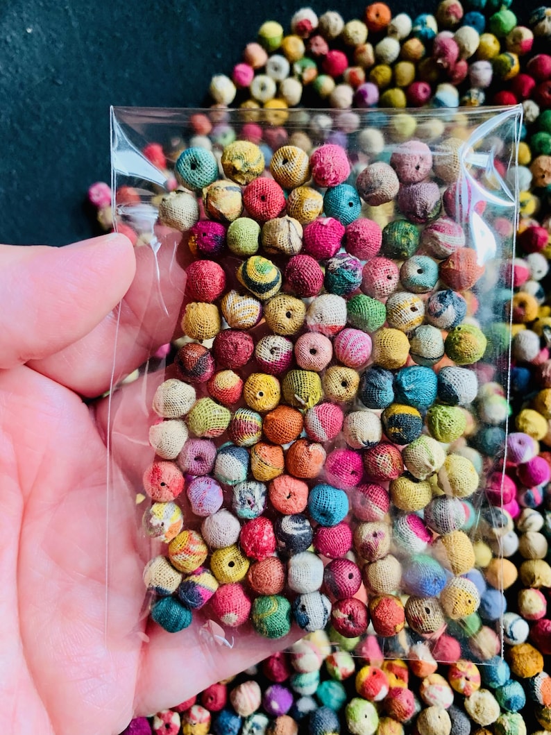 May include: A clear plastic bag filled with a variety of small, colorful fabric-covered beads. The beads are in shades of pink, yellow, green, blue, and red. The background is filled with more of the same beads, creating a vibrant and textured display.