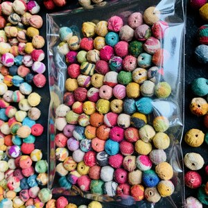 May include: A clear plastic bag filled with colorful, round beads. The beads are wrapped in a textured fabric and come in a variety of colors, including pink, blue, yellow, and green. The beads are scattered around the bag on a black surface.