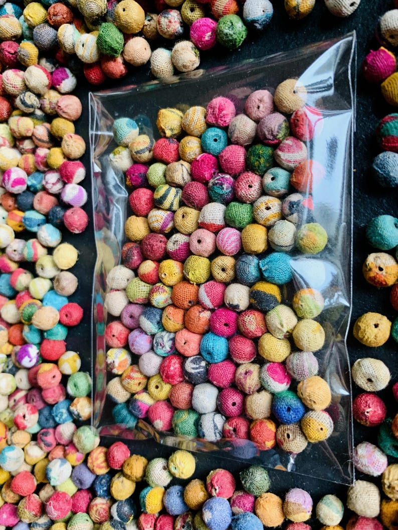 May include: A clear plastic bag filled with colorful, round beads. The beads are wrapped in a textured fabric and come in a variety of colors, including pink, blue, yellow, and green. The beads are scattered around the bag on a black surface.