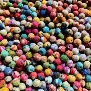 May include: A close-up shot of a collection of small, colorful, round objects. The objects are wrapped in various colors of thread, including red, blue, yellow, green, and pink. The texture of the thread is visible, creating a tactile appearance. The objects are densely packed together.