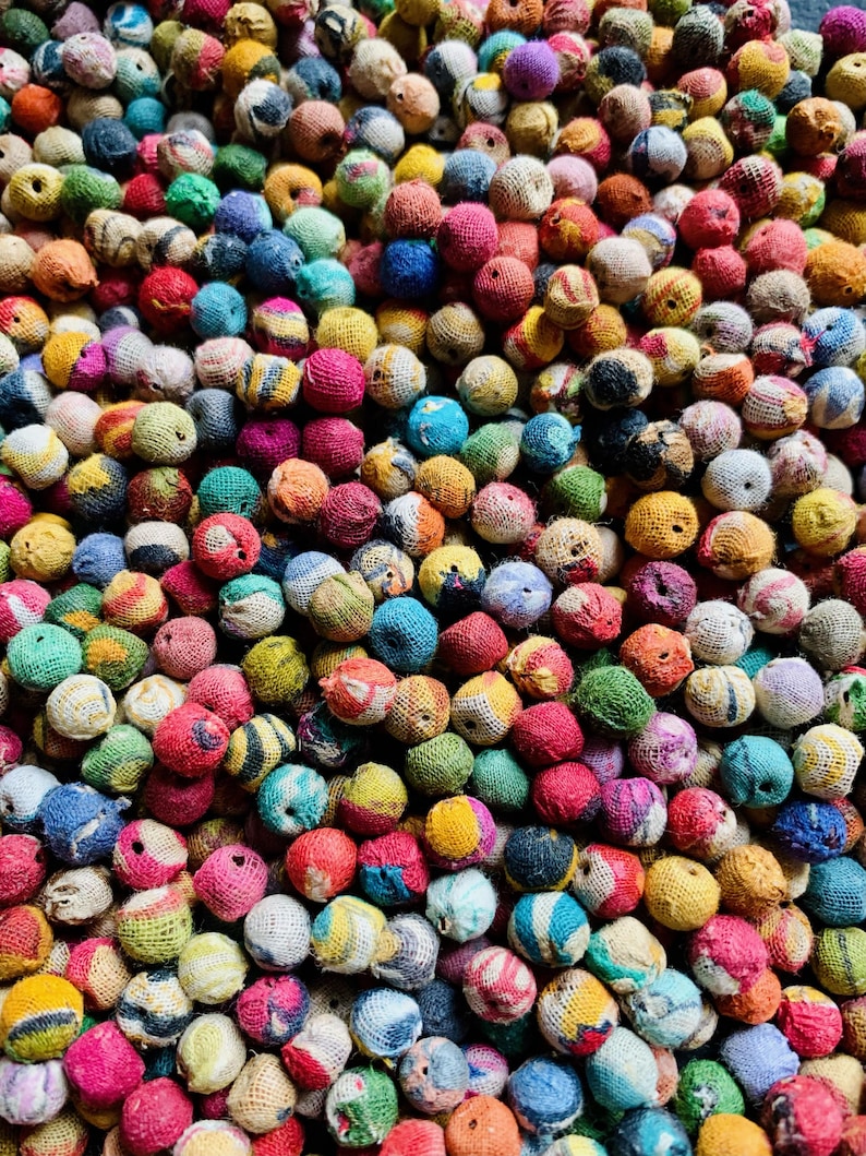 May include: A close-up shot of a collection of small, colorful, round objects. The objects are wrapped in various colors of thread, including red, blue, yellow, green, and pink. The texture of the thread is visible, creating a tactile appearance. The objects are densely packed together.