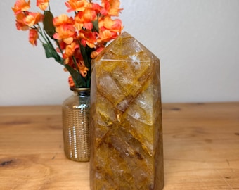 May include: A golden-yellow quartz crystal obelisk with a faceted top. The crystal sits on a wooden surface, with a small gold vase and orange flowers in the background. The crystal is approximately 6 inches tall.
