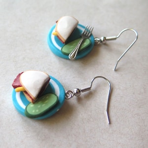 Pode incluir: Um par de brincos com um sanduíche em miniatura num prato azul com um picles e um garfo. Os brincos são de prata e têm um fecho de gancho.