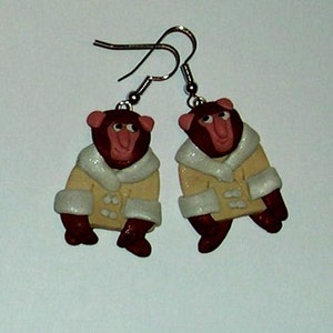 May include: A pair of earrings featuring brown monkey figures wearing cream-colored coats with white fur trim. The monkeys have pink noses and are wearing silver hooks.