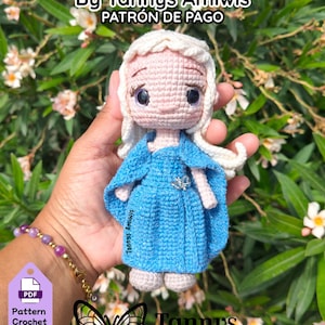 May include: A crocheted Daenerys doll in a blue dress with white hair, a silver brooch, and large eyes. The doll is held in a hand against a background of green foliage. Text on the image reads "DAENERYS By Tannys Amiwis PATRÓN DE PAGO".