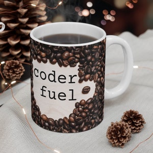Puede incluir: Taza de cerámica blanca con un diseño de granos de café marrones. La taza tiene el texto "coder fuel" en letras negras.