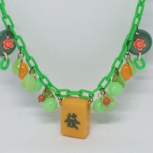 Op de afbeelding: Een groene plastic ketting met een gele mahjong-tegel als hanger. De ketting is versierd met groene, oranje en roze kralen.