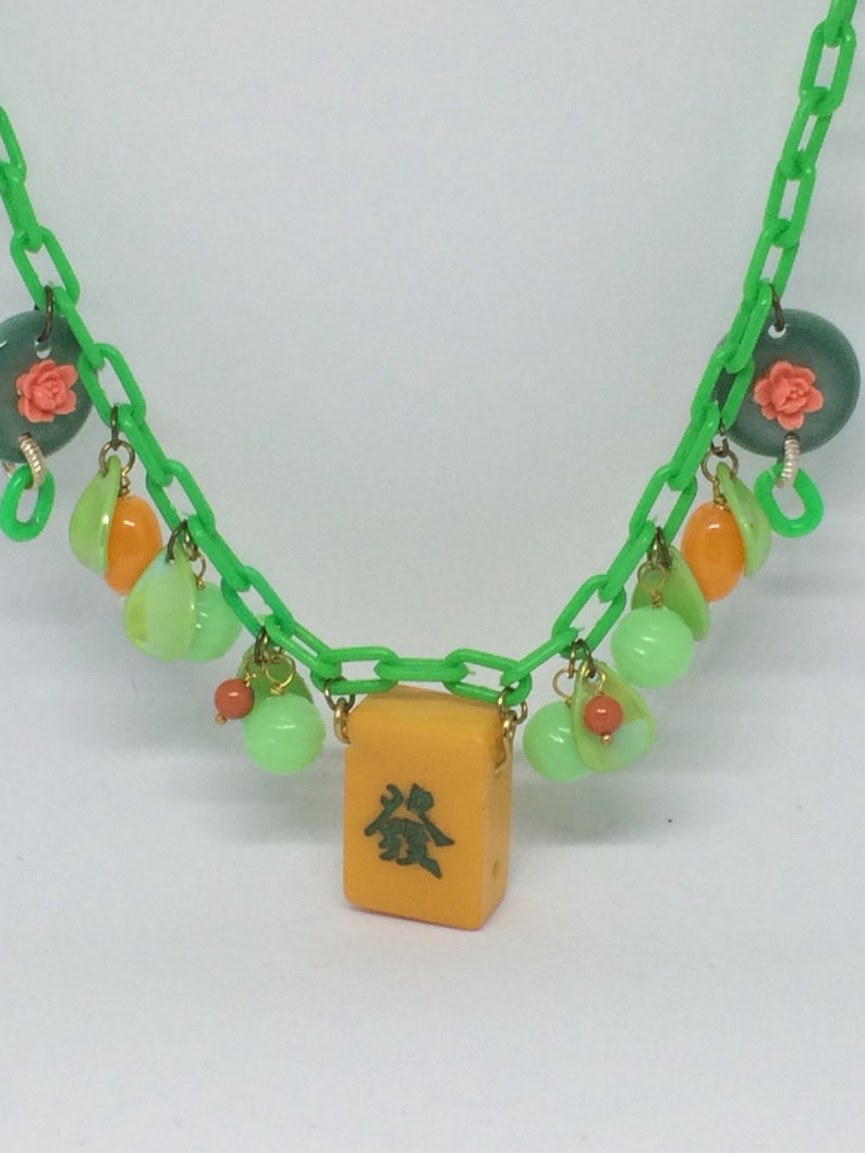 Op de afbeelding: Een groene plastic ketting met een gele mahjong-tegel als hanger. De ketting is versierd met groene, oranje en roze kralen.