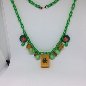 Op de afbeelding: Een groene plastic ketting met groene, oranje en roze kralen en een gele tegel met Chinese karakters.