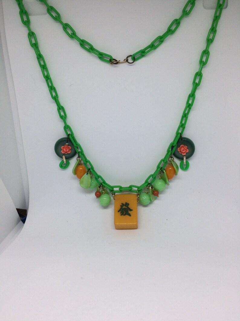 Op de afbeelding: Een groene plastic ketting met groene, oranje en roze kralen en een gele tegel met Chinese karakters.