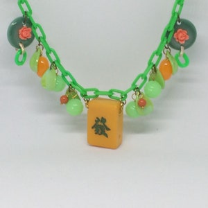 Op de afbeelding: Een groene plastic ketting met oranje en groene kralen, een groene en roze bloemenhanger en een gele mahjong-tegel met groene karakters. De tegel heeft het Chinese karakter voor "thee" erop.