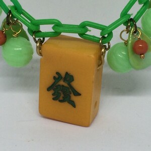 Op de afbeelding: Een groene plastic ketting met een gele mahjong-tegel hanger. De tegel heeft groene Chinese karakters erop. De ketting heeft ook groene en oranje kralen.