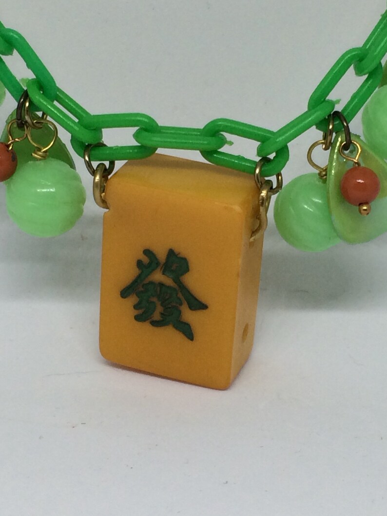 Op de afbeelding: Een groene plastic ketting met een gele mahjong-tegel hanger. De tegel heeft groene Chinese karakters erop. De ketting heeft ook groene en oranje kralen.