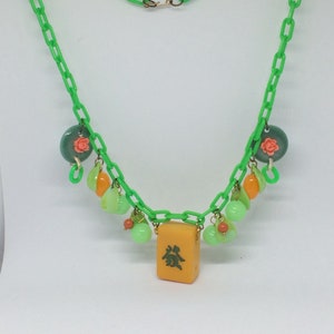 Op de afbeelding: Een groene plastic ketting met een gele mahjong-tegel als hanger. De ketting is versierd met groene, oranje en roze glazen kralen.