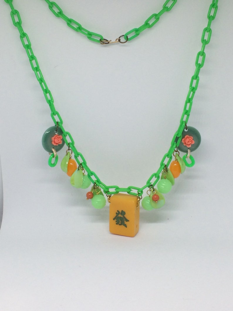 Op de afbeelding: Een groene plastic ketting met een gele mahjong-tegel als hanger. De ketting is versierd met groene, oranje en roze glazen kralen.