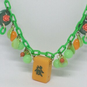 Op de afbeelding: Een groene plastic ketting met verschillende bedels, waaronder groene en oranje kralen, een groene en roze bloemenbedel en een gele mahjong-tegel met Chinese karakters.