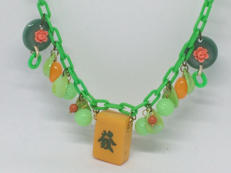 Op de afbeelding: Een groene plastic ketting met verschillende bedels, waaronder groene en oranje kralen, een groene en roze bloemenbedel en een gele mahjong-tegel met Chinese karakters.