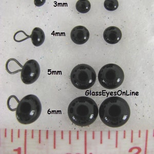May include: Black glass eyes with wire loops in various sizes, ranging from 3mm to 6mm. The eyes are displayed on a white surface with a ruler for size reference. The text "GlassEyesOnLine" is visible in the image.