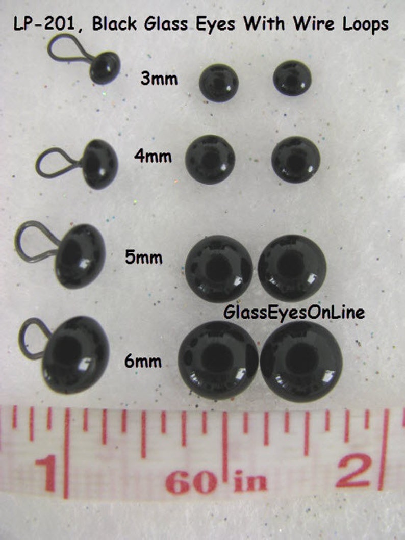 May include: Black glass eyes with wire loops in various sizes, ranging from 3mm to 6mm. The eyes are displayed on a white surface with a ruler for size reference. The text "GlassEyesOnLine" is visible in the image.