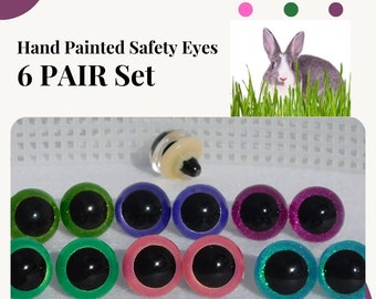 May include: Six pairs of hand-painted safety eyes for crafts. The eyes are in various colors, including green, blue, pink, purple, and teal. Each eye has a black pupil and a glitter finish. The set also includes a pair of plastic safety eyes.