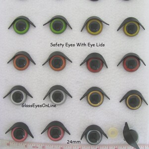 Puede incluir: Surtido de ojos de seguridad de 24 mm con p&aacute;rpados en varios colores, incluidos azul, verde, naranja, rojo, negro, blanco y dorado. Los ojos est&aacute;n dispuestos en filas sobre una superficie blanca con una regla para la escala. El texto "Safety Eyes With Eye Lids" y "GlassEyesOnLine" es visible en la imagen.