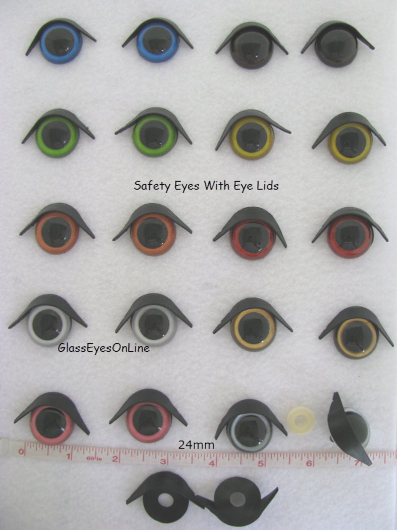 Puede incluir: Surtido de ojos de seguridad de 24 mm con p&aacute;rpados en varios colores, incluidos azul, verde, naranja, rojo, negro, blanco y dorado. Los ojos est&aacute;n dispuestos en filas sobre una superficie blanca con una regla para la escala. El texto "Safety Eyes With Eye Lids" y "GlassEyesOnLine" es visible en la imagen.