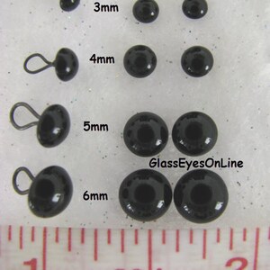 May include: Black glass eyes with wire loops in sizes 3mm, 4mm, 5mm, and 6mm. The eyes are shown on a white surface with a ruler for scale. The text "GlassEyesOnLine" is visible in the image.