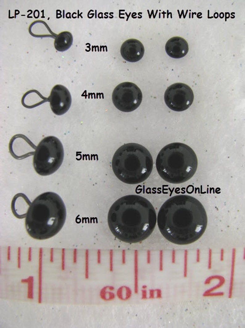 May include: Black glass eyes with wire loops in sizes 3mm, 4mm, 5mm, and 6mm. The eyes are shown on a white surface with a ruler for scale. The text "GlassEyesOnLine" is visible in the image.