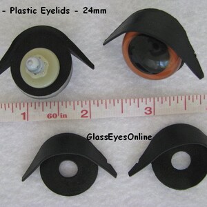 May include: Four black plastic eyelids for doll eyes, measuring 24mm in diameter. The eyelids are shown on a white surface with a ruler for scale. The text '#El-1 - Plastic Eyelids - 24mm' and 'GlassEyesOnline' are visible in the image.