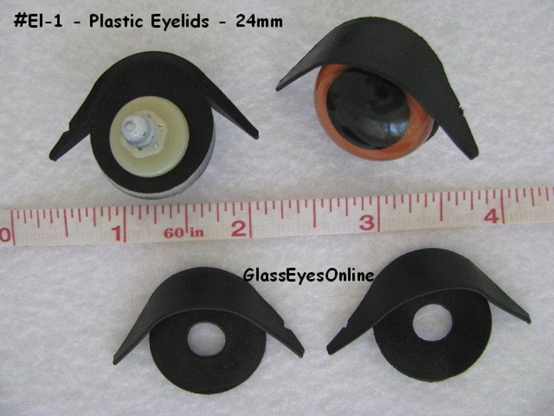 May include: Four black plastic eyelids for doll eyes, measuring 24mm in diameter. The eyelids are shown on a white surface with a ruler for scale. The text '#El-1 - Plastic Eyelids - 24mm' and 'GlassEyesOnline' are visible in the image.